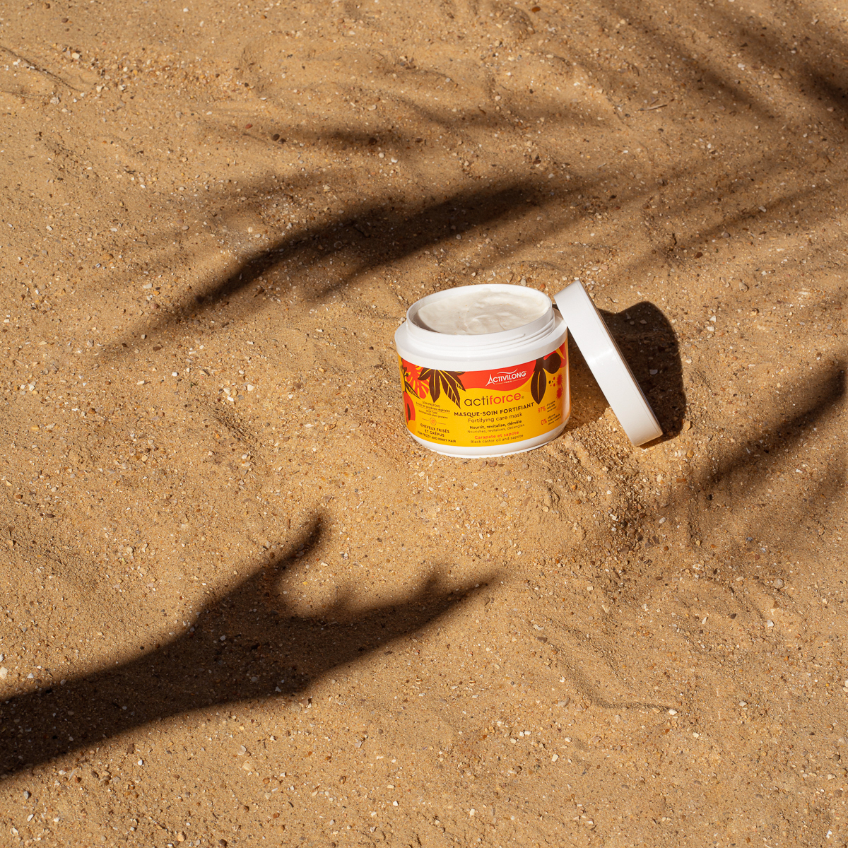 Produit capillaire posé sur le sable entre l'ombre d'un palmier et d'une main tendue